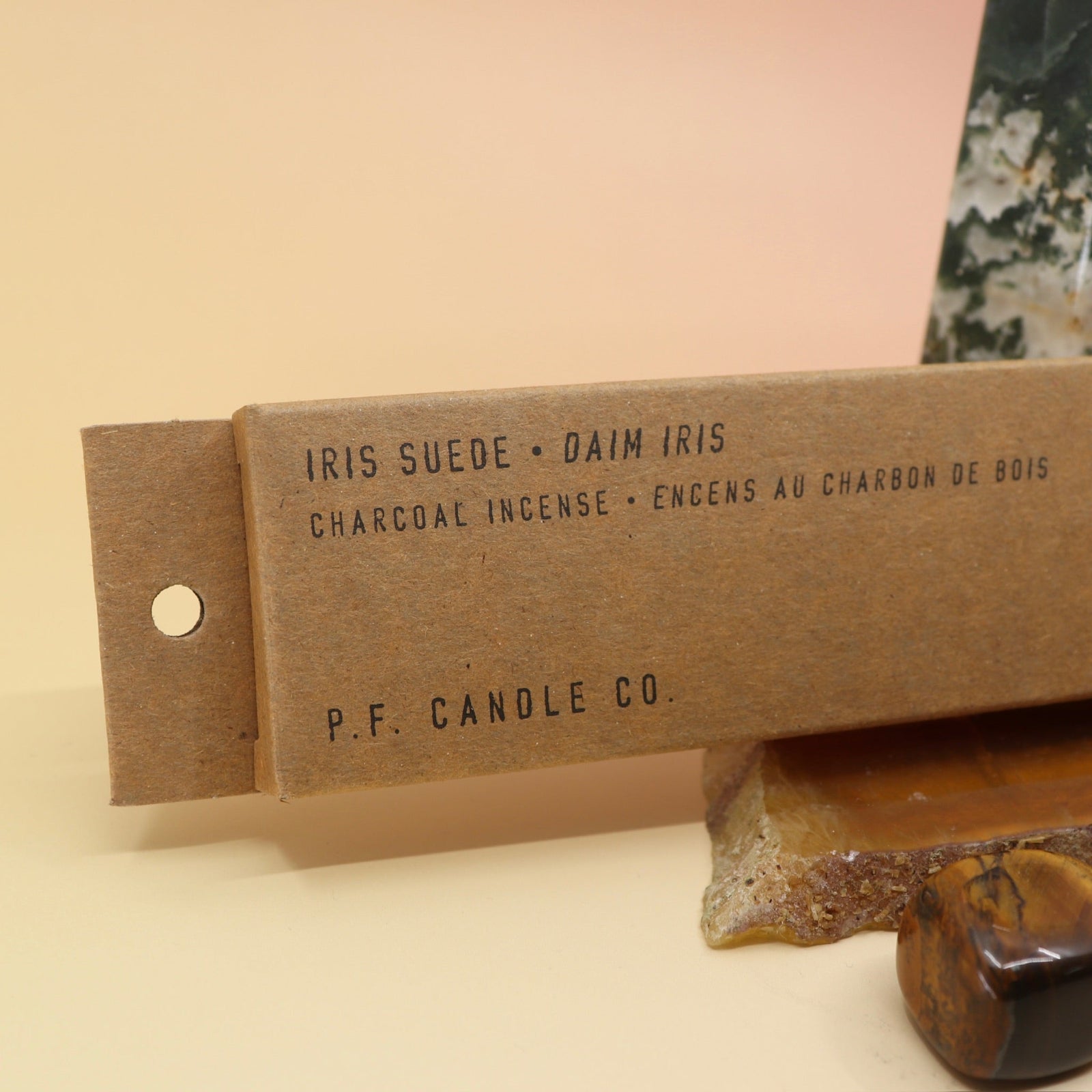 The P.F. Candle Co. Iris Suede Incense on an agate slice in front of a crystal tower all on a pink and yellow background.