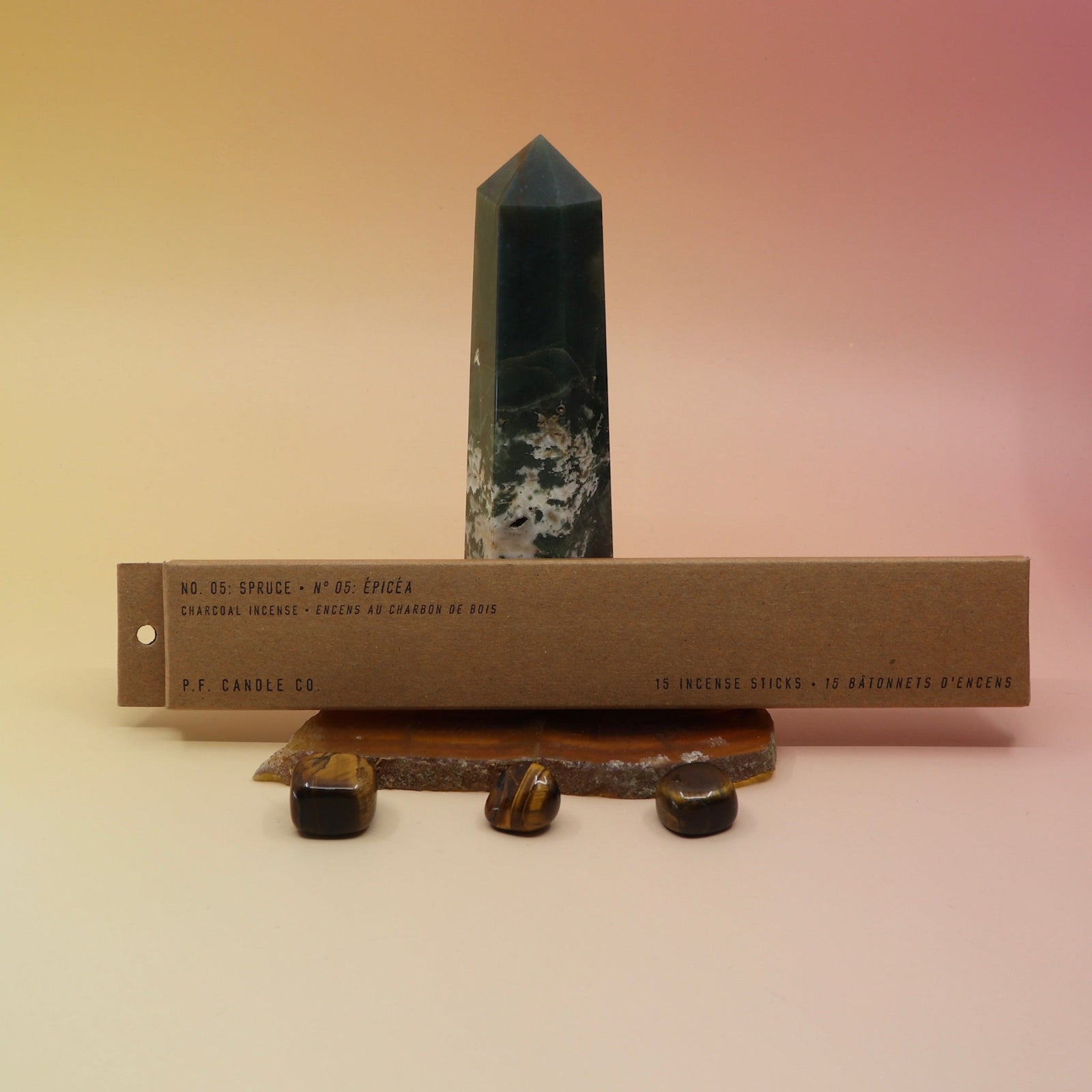 The P.F. Candle Co. Spruce Incense on an agate slice and in front of a crystal tower all on a pink and yellow background.