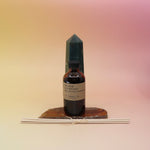 The Iris Suede Reed Diffuser on an agate slice in front of a crystal tower, all on a pink and yellow background.