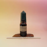 The Iris Suede Reed Diffuser on an agate slice in front of a crystal tower, all on a pink and yellow background.