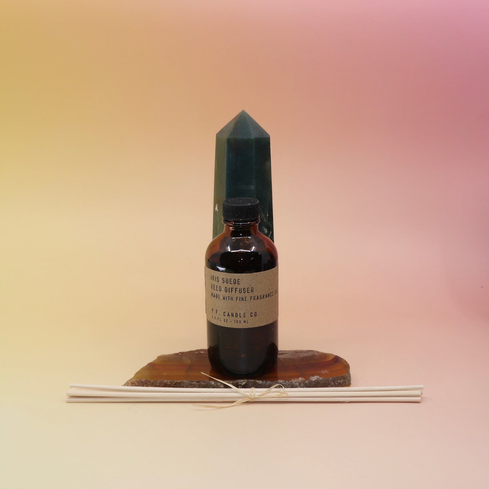 The Iris Suede Reed Diffuser on an agate slice in front of a crystal tower, all on a pink and yellow background.