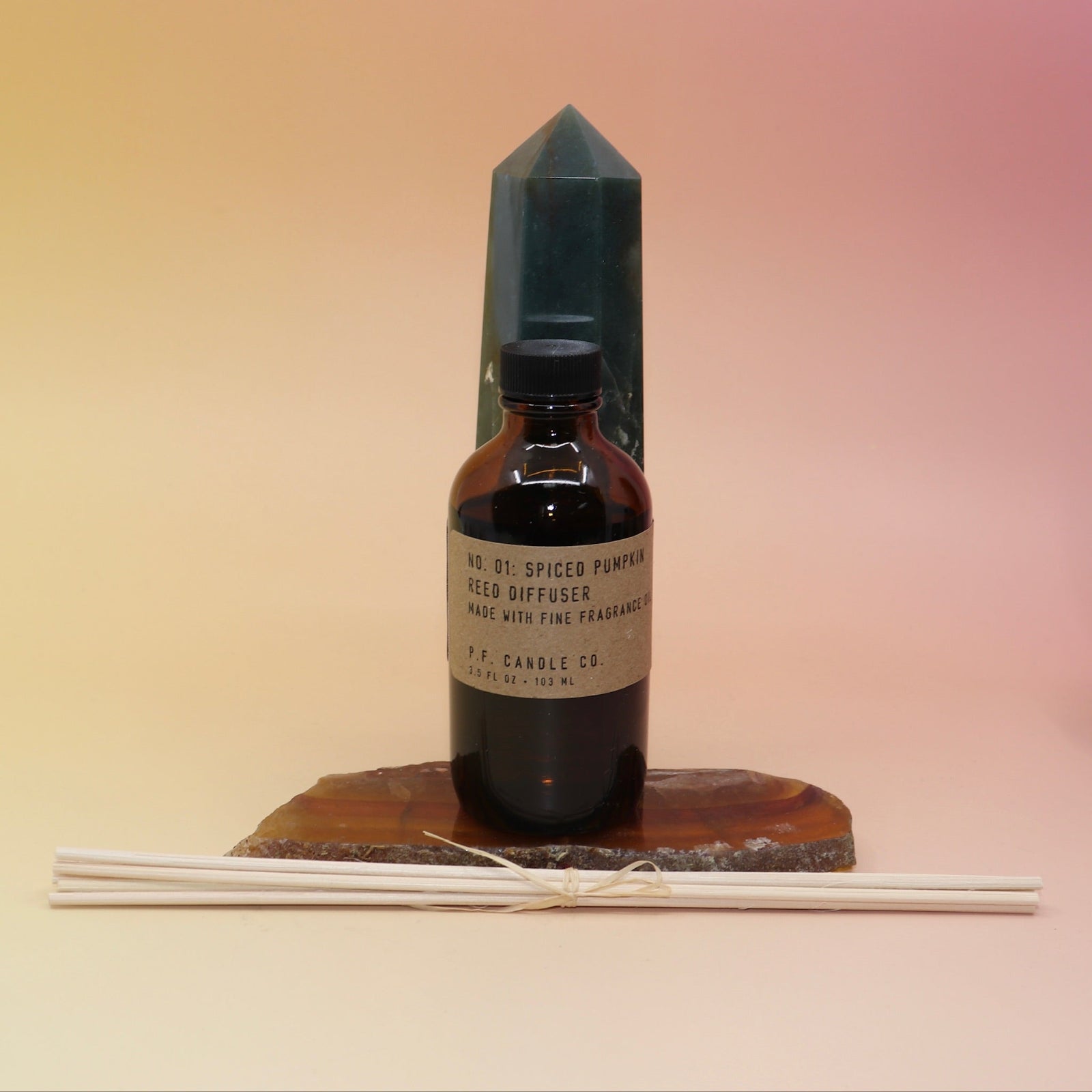 The P.F. Candle Co. Spiced Pumpkin - 3.5 oz Reed Diffuser on an agate slice in front of a crystal tower all on a pink and yellow background.