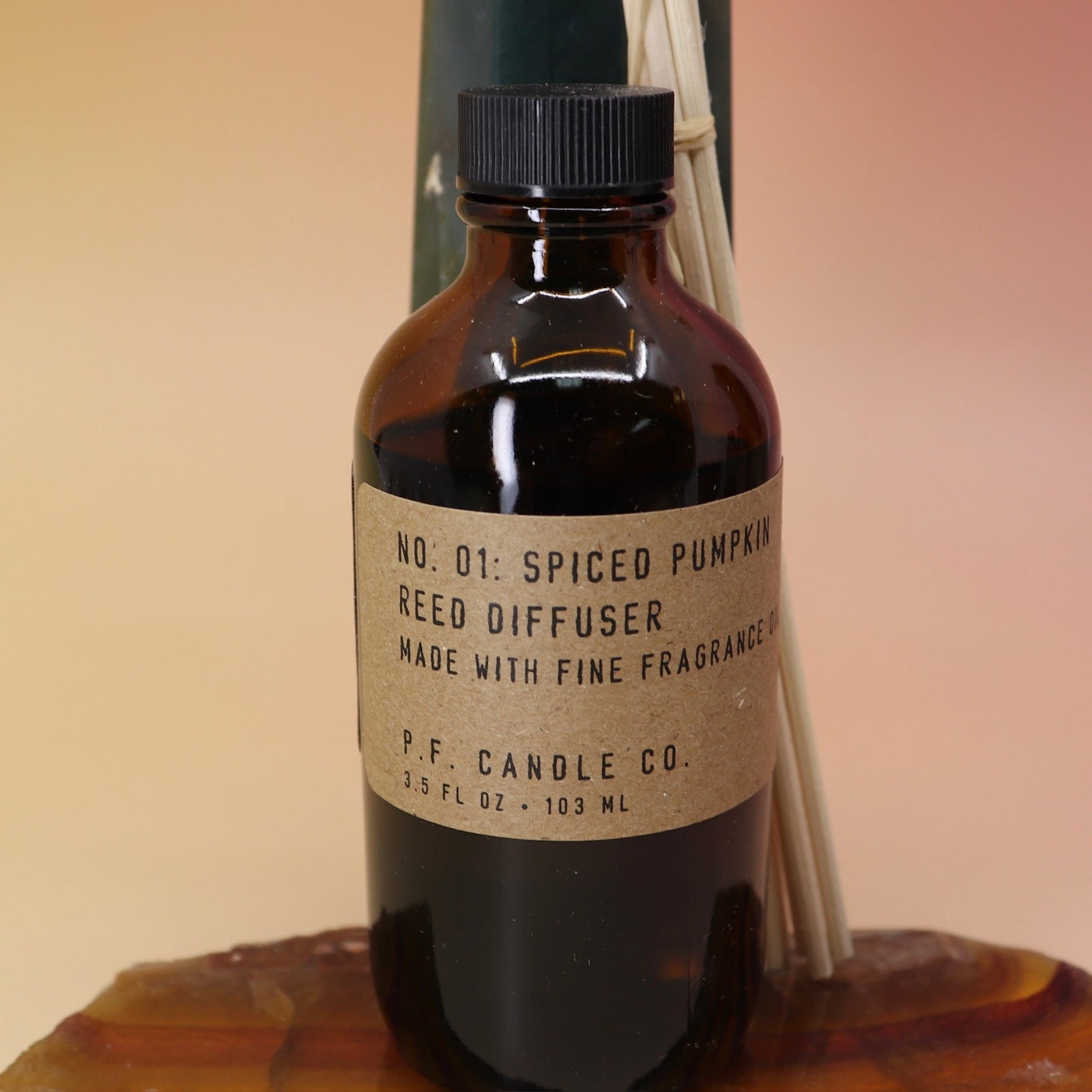 An up-close image of the P.F. Candle Co. Spiced Pumpkin - 3.5 oz Reed Diffuser on an agate slice in front of a crystal tower all on a pink and yellow background.