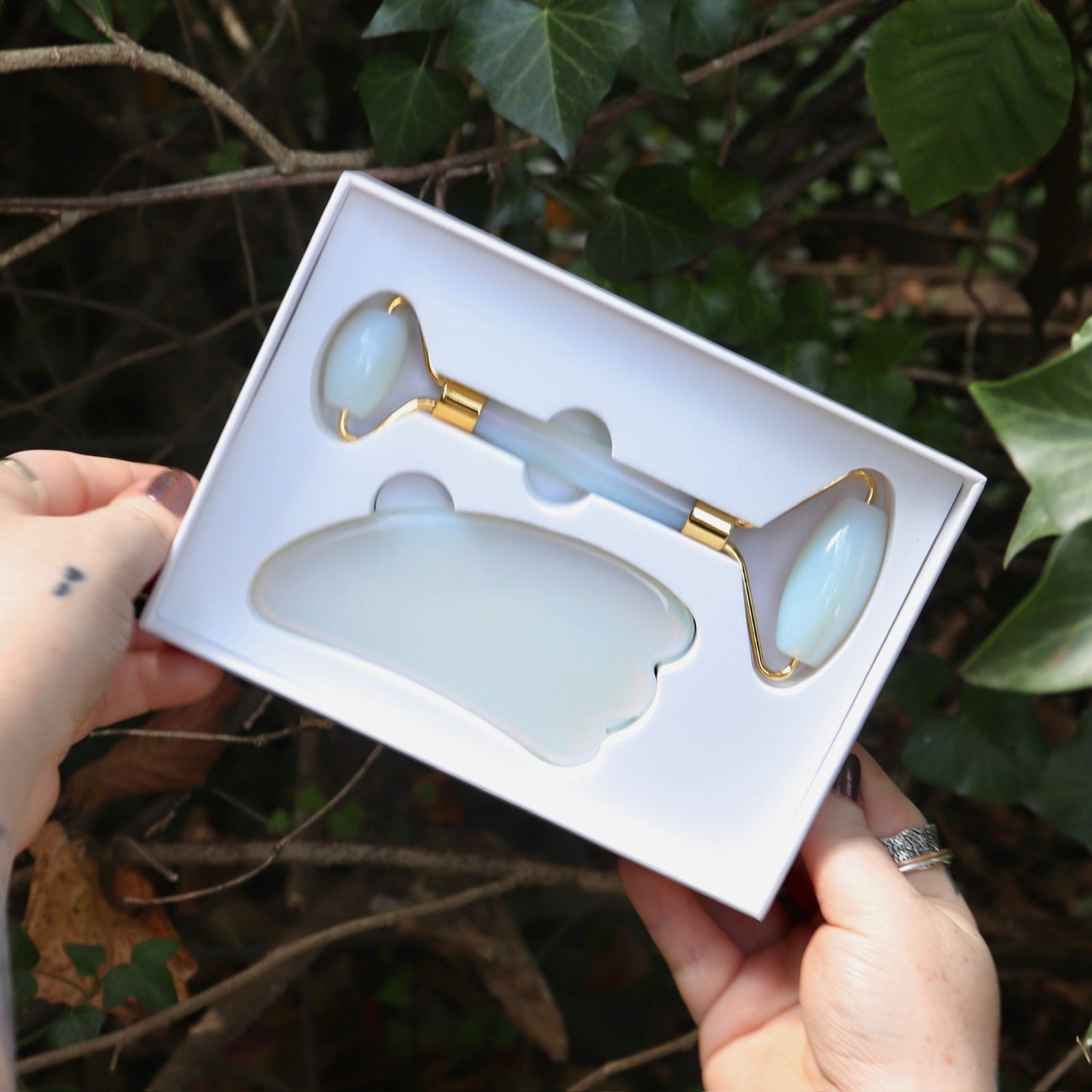 Someone holding the Opalite Crystal Facial Roller Set in front of a green bush.