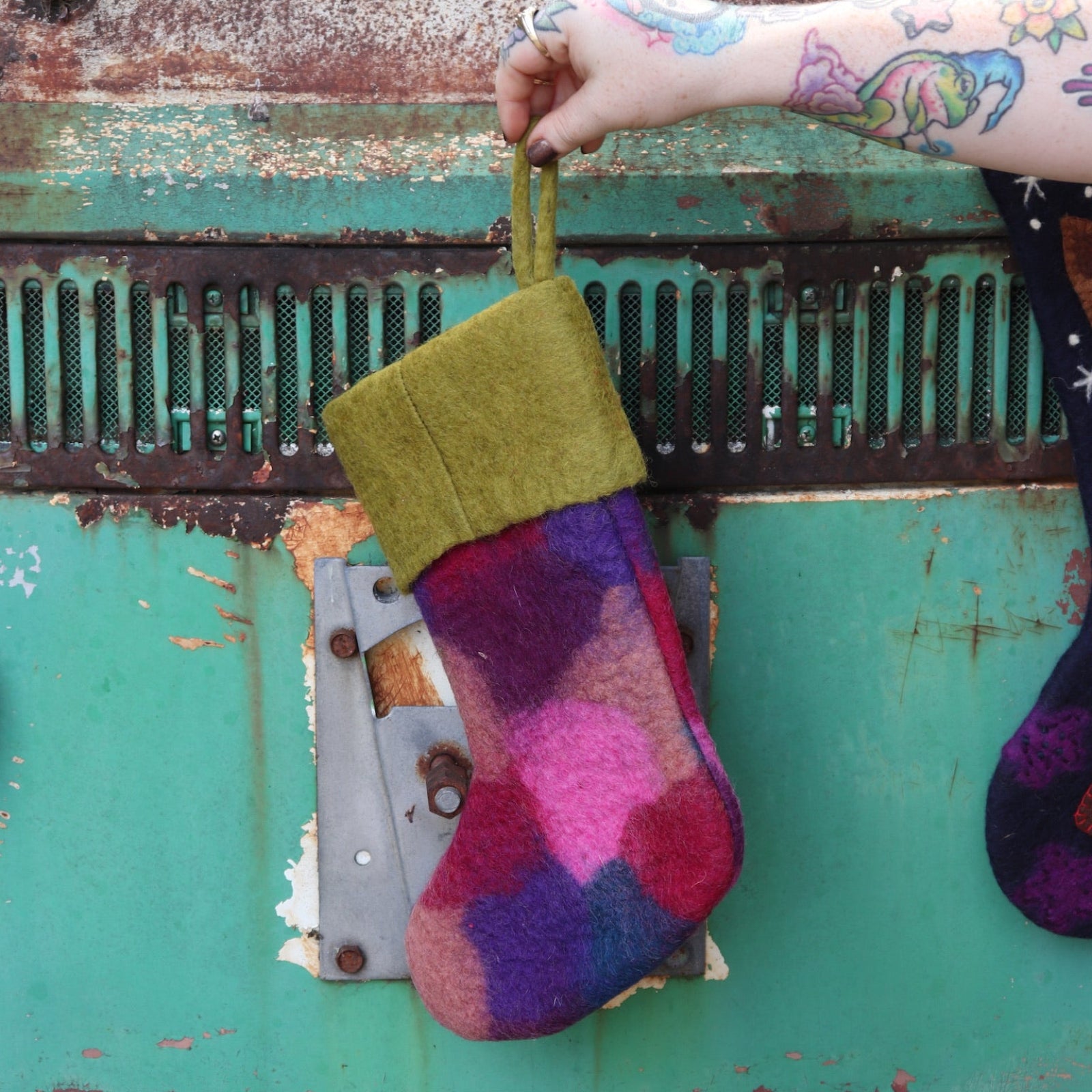 Someone holding the olive Tie-Dye Peace Pure Wool Felt Handcrafted Stocking in front of a vintage teal Volkswagen bus, showing off the back.