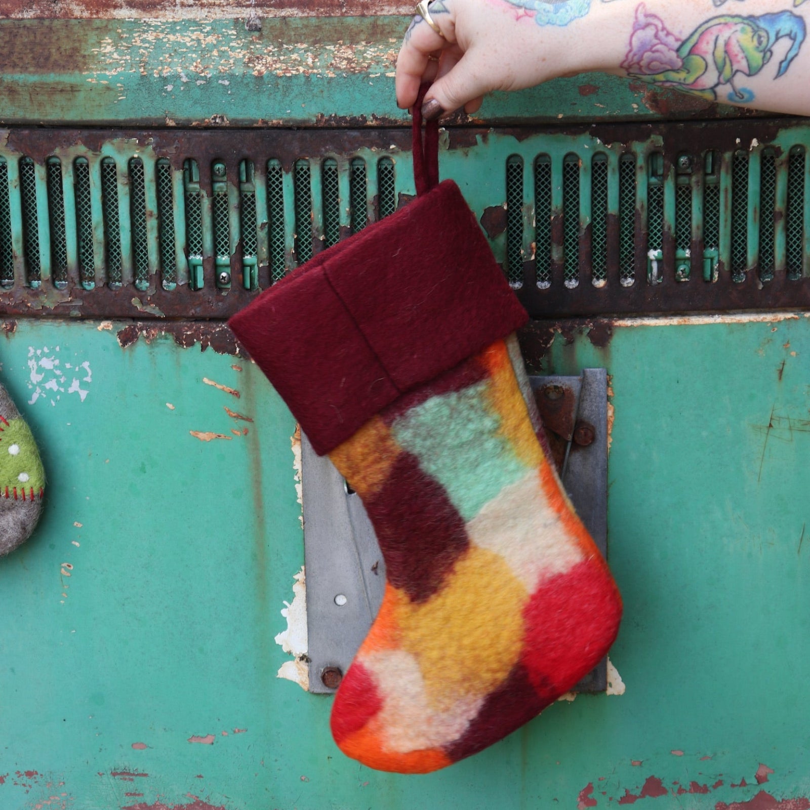 Someone holding the burgundy Tie-Dye Peace Pure Wool Felt Handcrafted Stocking in front of a vintage teal Volkswagen bus, showing off the back.