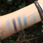 Someone wearing test swabs of the Healing Waters 6 Pan Vegan Eyeshadow Palette on their arm in front of a green bush.