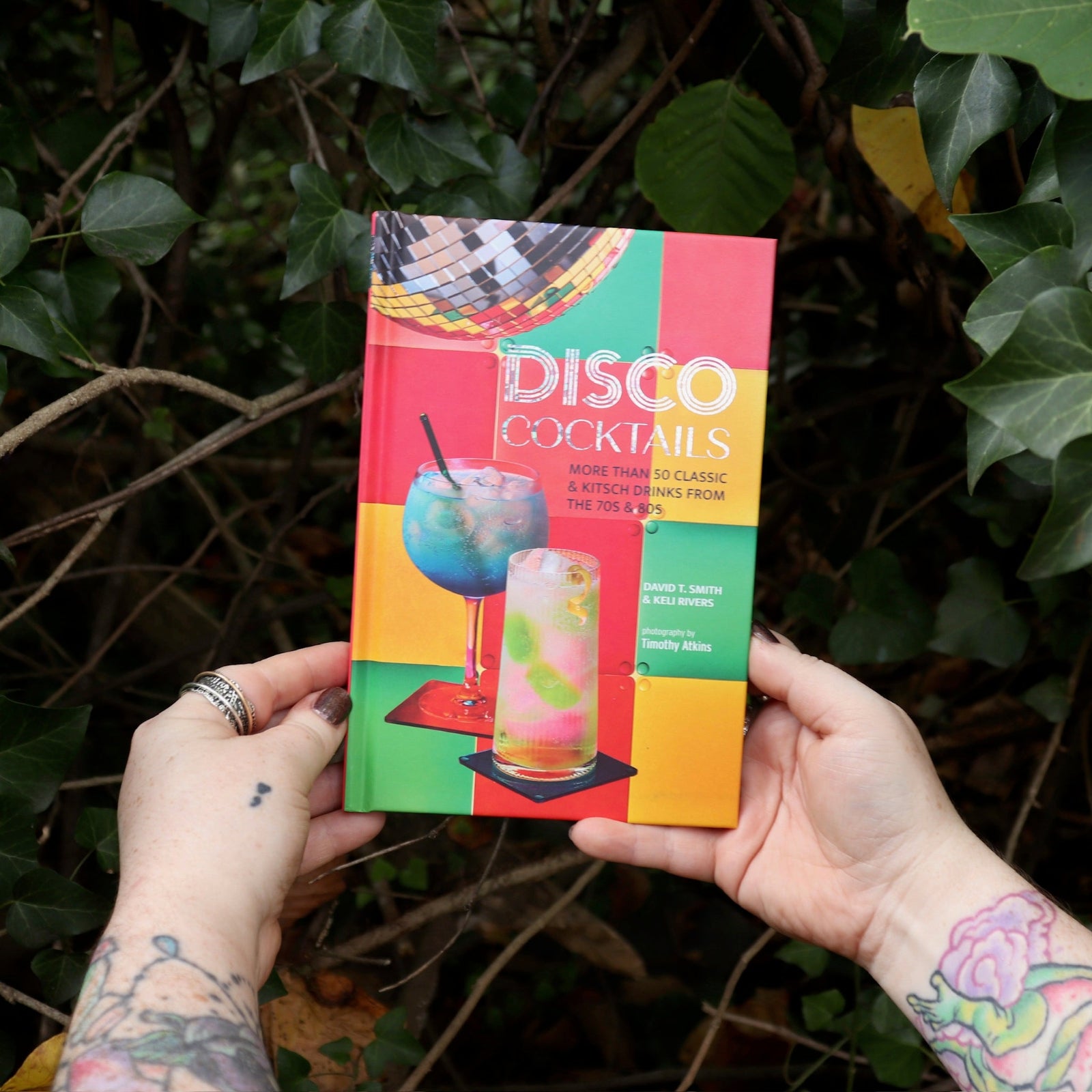 Someone holding the Disco Cocktails Hardcover Book in front of a green bush.