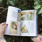 Someone holding the Sacred Herbs of Yule and Christmas open in front of a green bush, showing off the pages.
