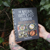 Someone holding the Natural Witch's Cookbook Hardcover Book in front of a green bush.