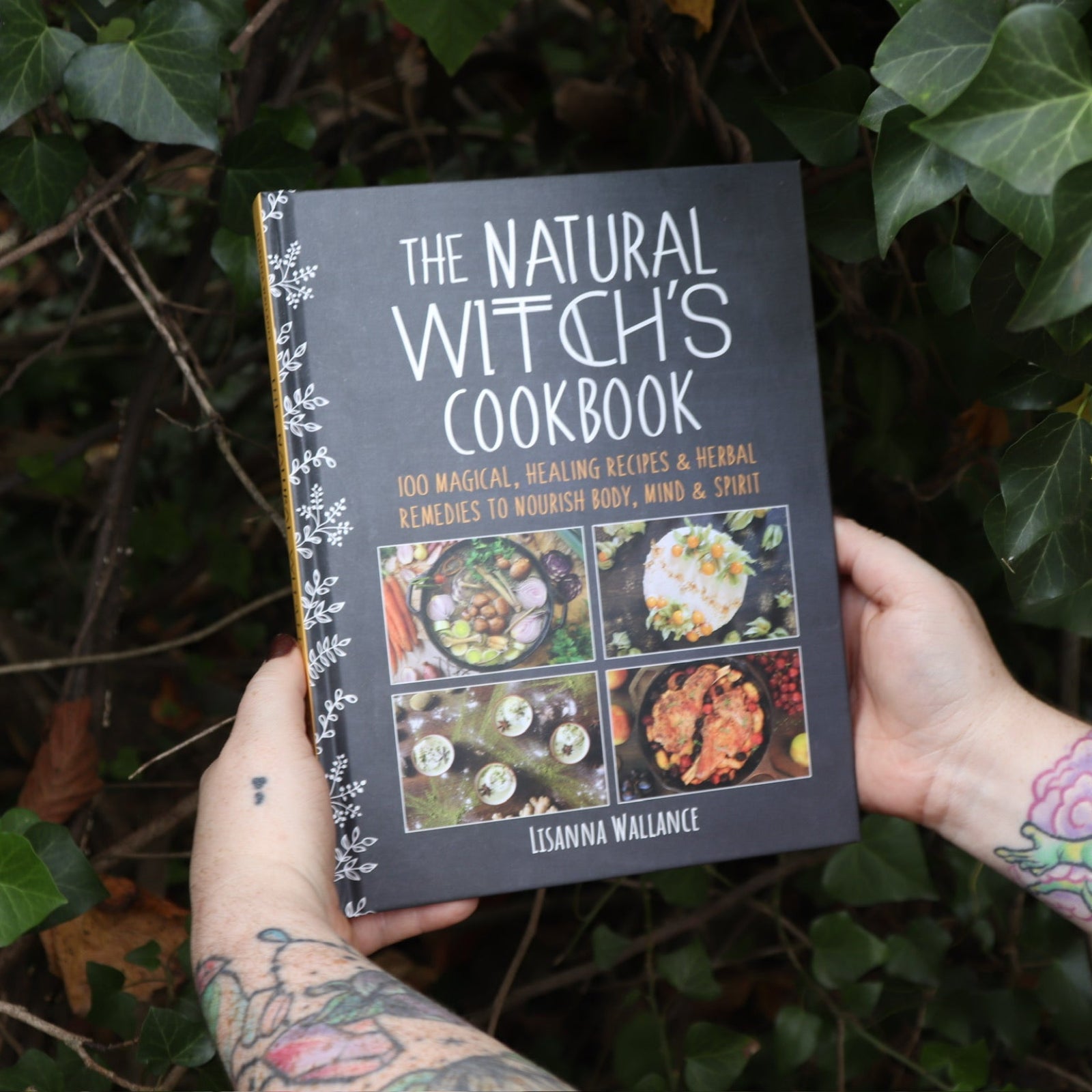 Someone holding the Natural Witch's Cookbook Hardcover Book in front of a green bush.