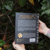 Someone holding the Natural Witch's Cookbook Hardcover Book in front of a green bush, showing off the back.
