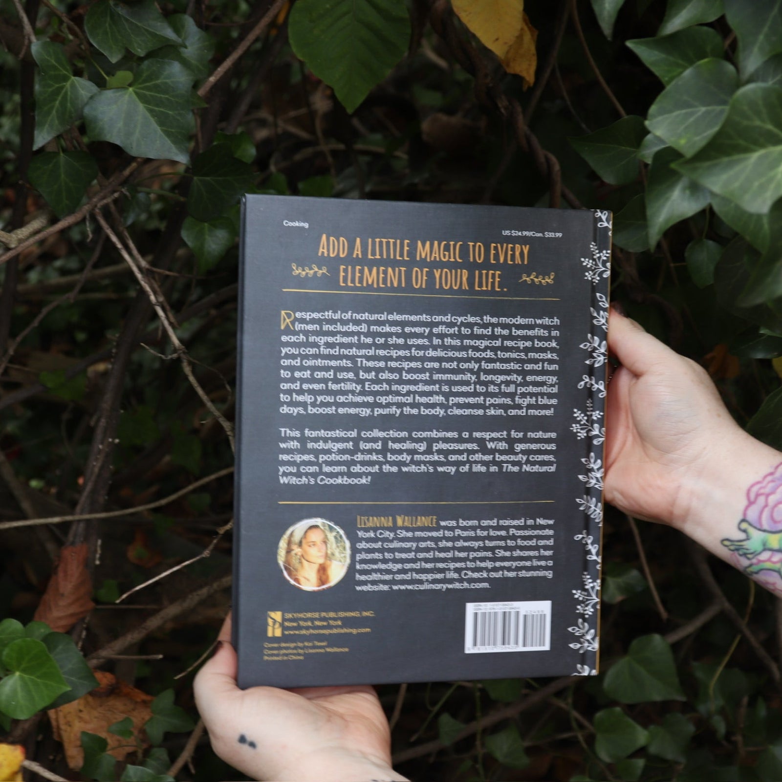 Someone holding the Natural Witch's Cookbook Hardcover Book in front of a green bush, showing off the back.