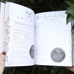 Someone holding the Natural Witch's Cookbook Hardcover Book open in front of a green bush, showing off the pages.