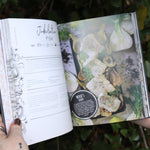 Someone holding the Natural Witch's Cookbook Hardcover Book open in front of a green bush, showing off the pages.