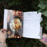 Someone holding the Natural Witch's Cookbook Hardcover Book open in front of a green bush, showing off the pages.