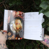 Someone holding the Natural Witch's Cookbook Hardcover Book open in front of a green bush, showing off the pages.