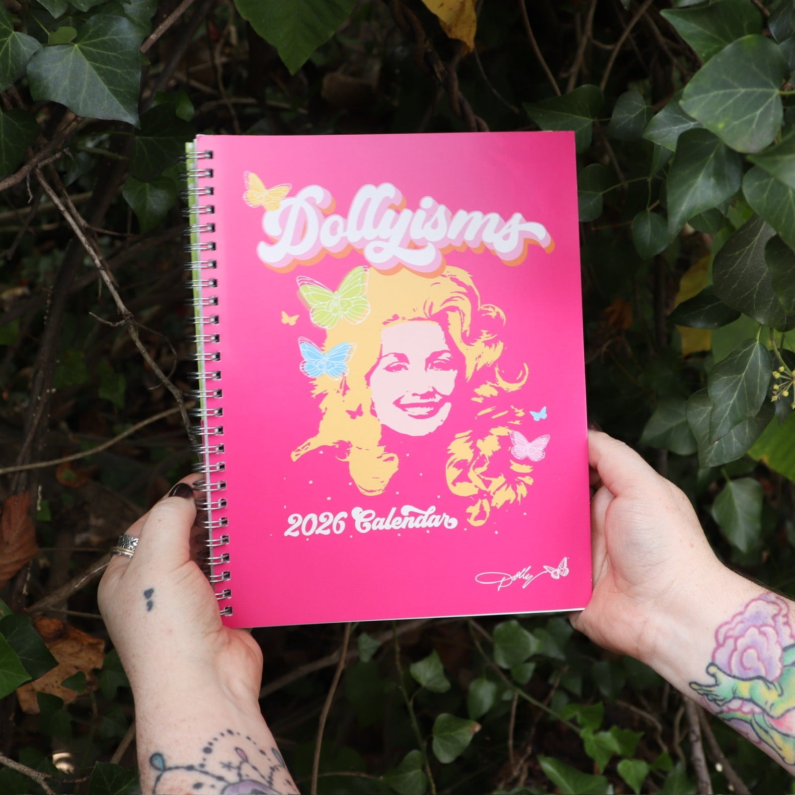 Someone holding the Dollyisms: Dolly Parton's 12-Month 2026 Softcover Planner in front of a green bush.