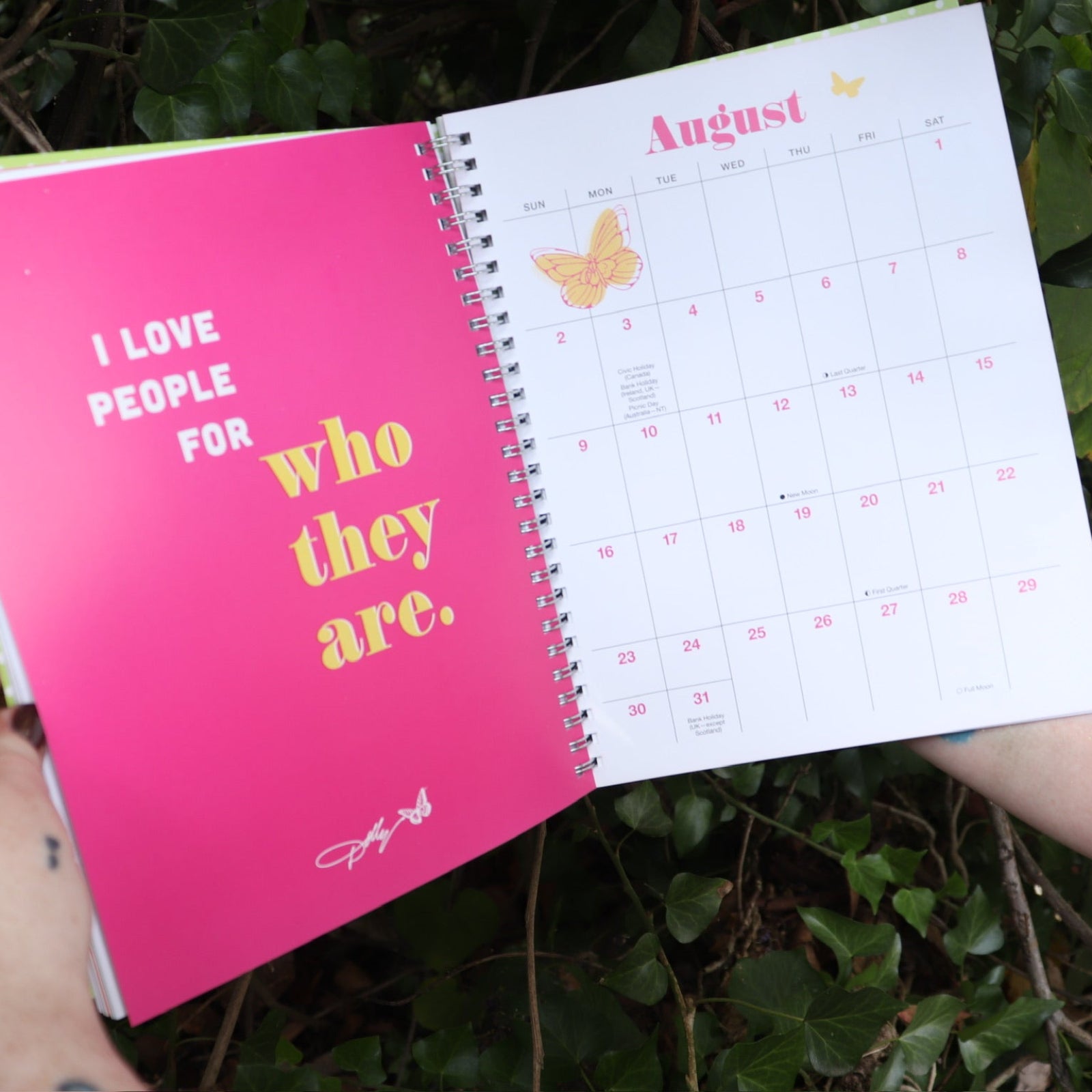 Someone holding the Dollyisms: Dolly Parton's 12-Month 2026 Softcover Planner open in front of a green bush, showing off the pages.