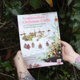 Someone holding the Scandinavian Christmas Crafts Paperback Book in front of a green bush.