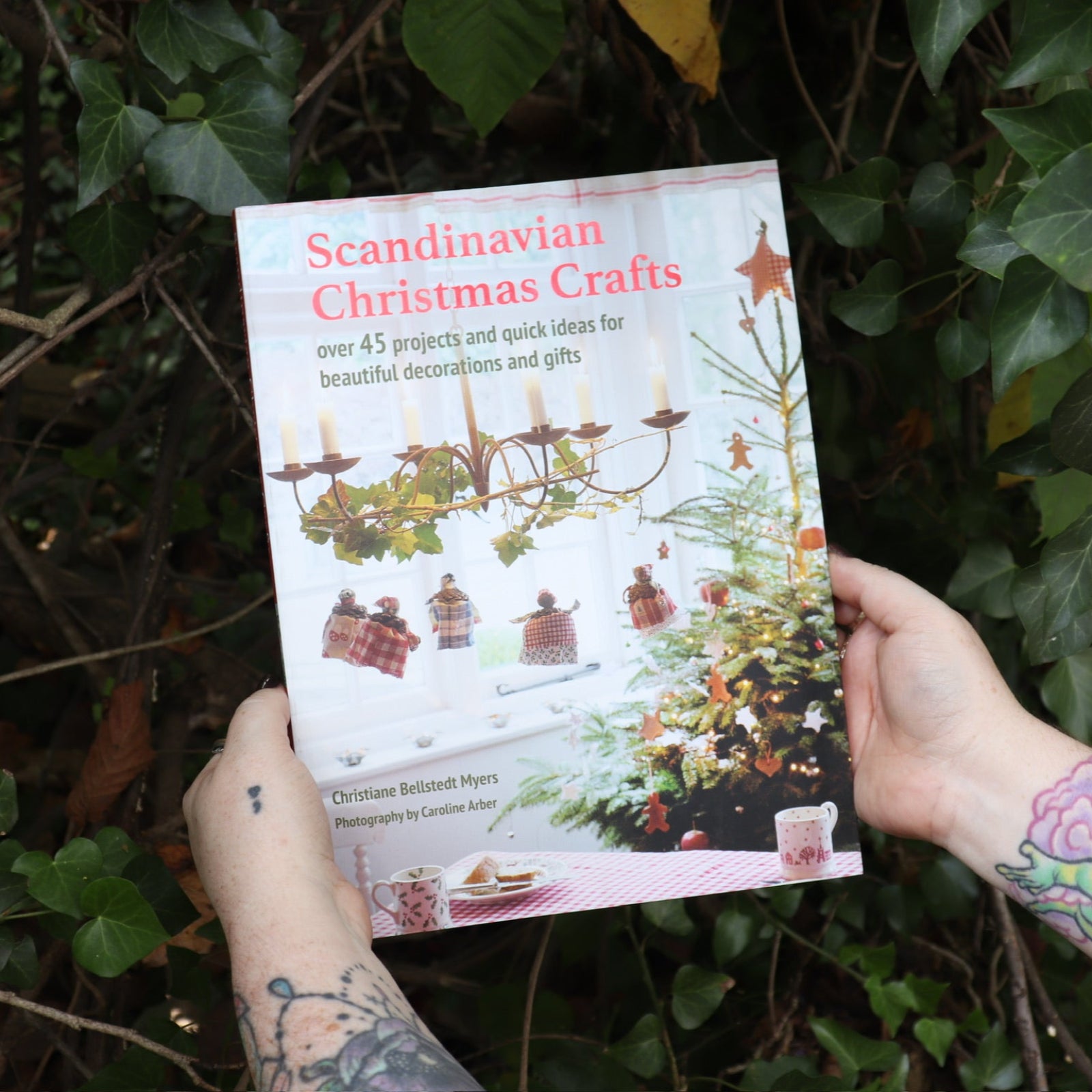 Someone holding the Scandinavian Christmas Crafts Paperback Book in front of a green bush.