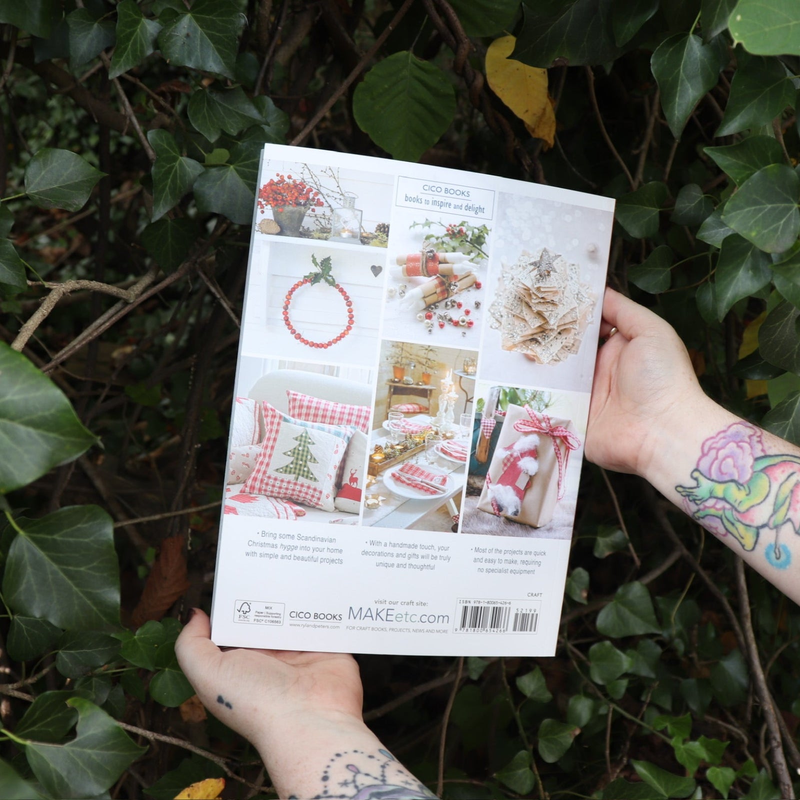Someone holding the Scandinavian Christmas Crafts Paperback Book in front of a green bush, showing off the back.