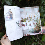 Someone holding the Scandinavian Christmas Crafts Paperback Book open in front of a green bush, showing off the pages.