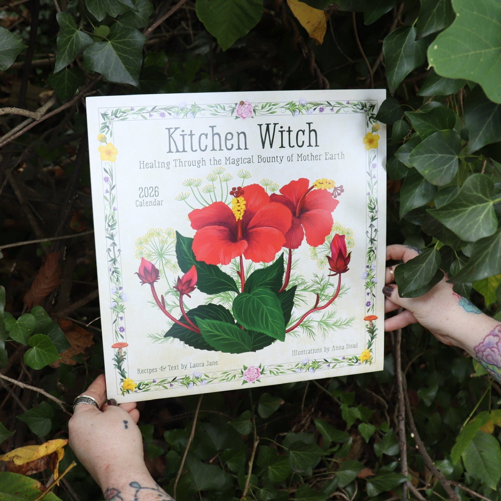 Someone holding the Kitchen Witch 2026 Wall Calendar in front of a green bush.