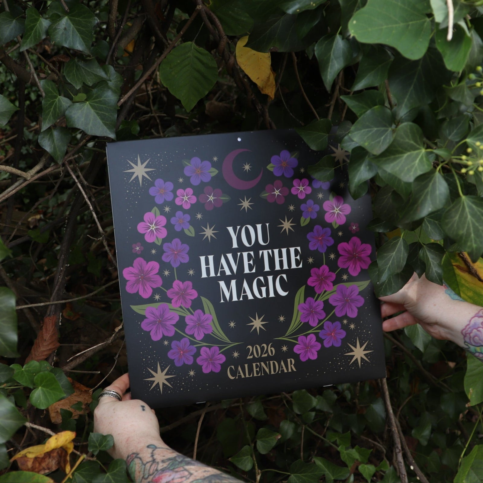 Someone holding the You Have the Magic 2026 Wall Calendar in front of a green bush.