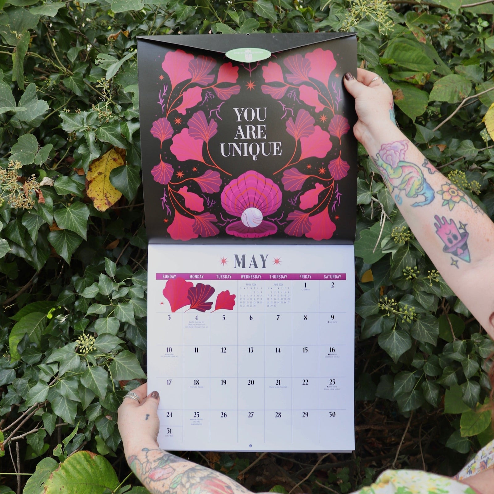 Someone holding the You Have the Magic 2026 Wall Calendar open in front of a green bush, showing off the pages.