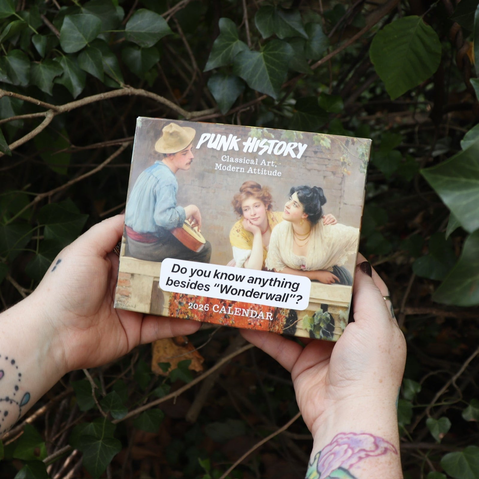 Someone holding the Punk History 2026 Day-to-Day Calendar in front of a green bush.