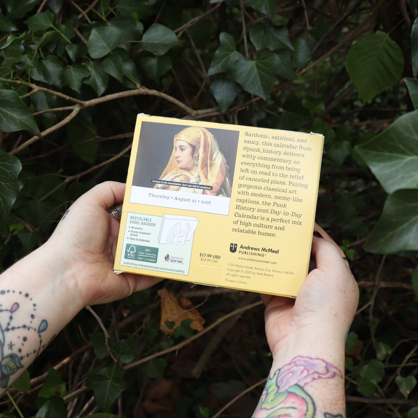 Someone holding the Punk History 2026 Day-to-Day Calendar in front of a green bush, showing off the back.