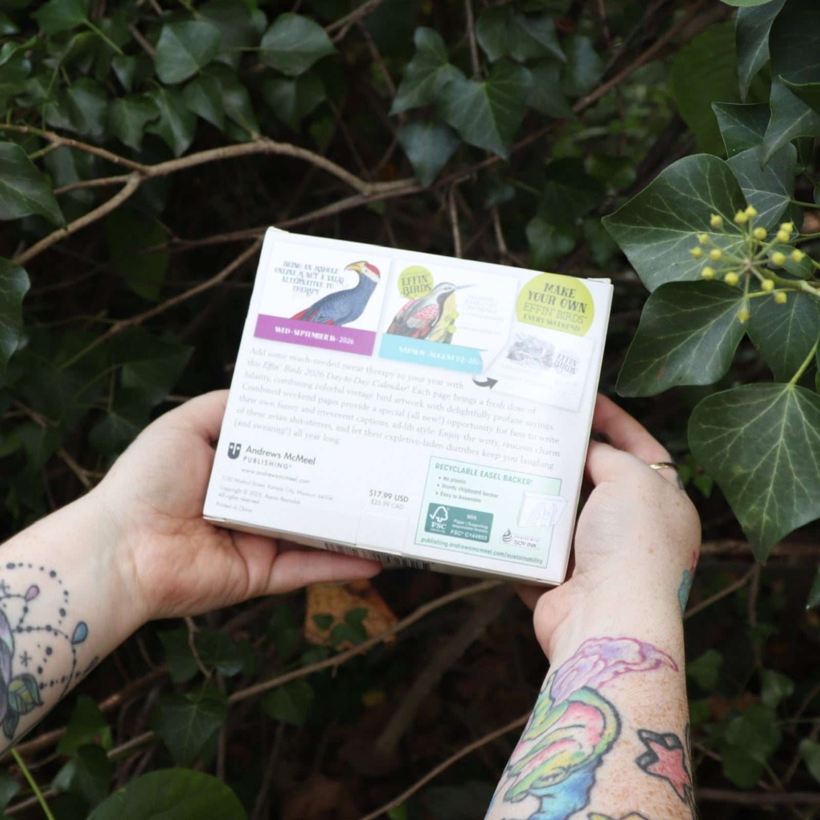 Someone holding the Effin' Birds 2026 Day-to-Day Calendar in front of a green bush, showing off the back.