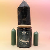 The Celestial Catskills 12oz Soy Candle in front of a large crystal tower and surrounded by two smaller crystals all on a pink and yellow background.