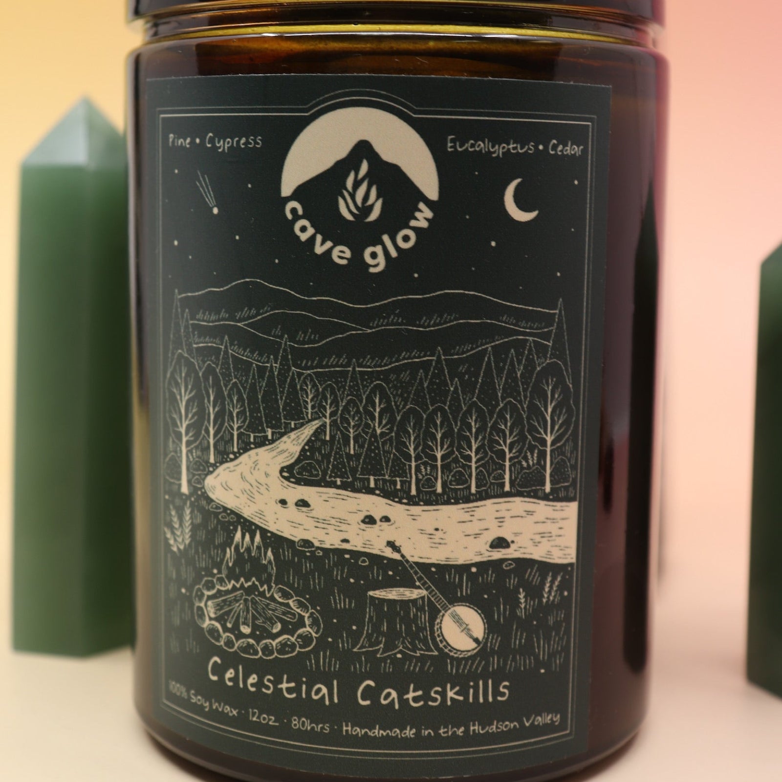 The Celestial Catskills 12oz Soy Candle in front of a large crystal tower and surrounded by two smaller crystals all on a pink and yellow background.
