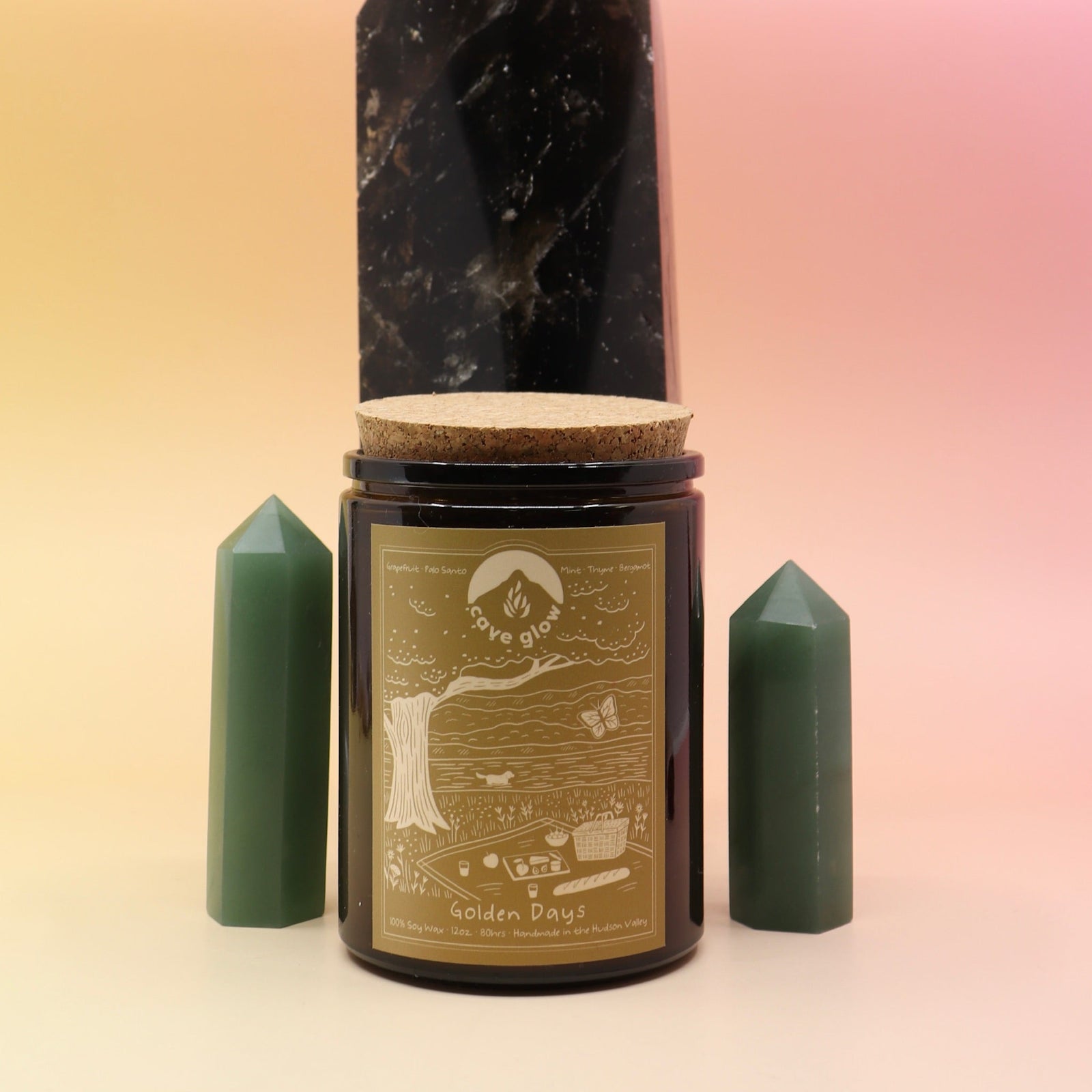 The Golden Days 12oz Soy Candle in front of a large crystal tower surrounded by two smaller crystal towers all on a pink and yellow background.