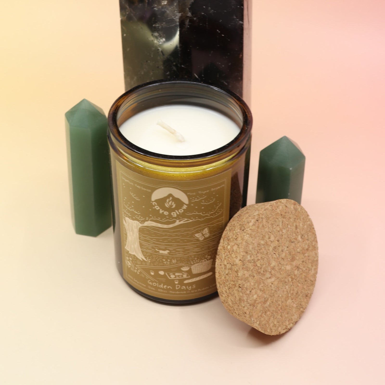 The Golden Days 12oz Soy Candle in front of a large crystal tower surrounded by two smaller crystal towers all on a pink and yellow background.