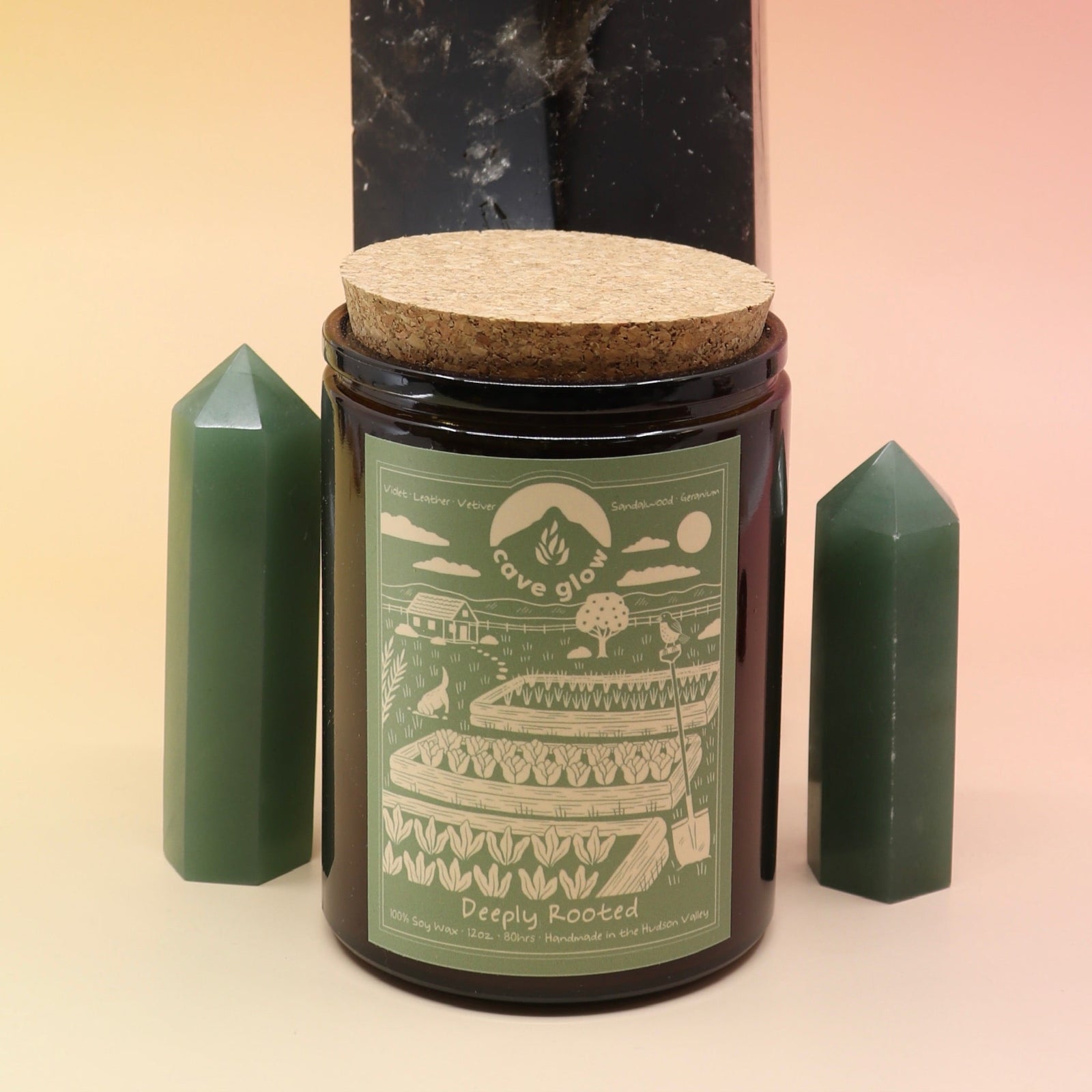 The Deeply Rooted 12oz Soy Candle in front of a large crystal tower surrounded by two smaller crystal towers all on a pink and yellow background.