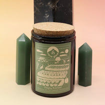 The Deeply Rooted 12oz Soy Candle in front of a large crystal tower surrounded by two smaller crystal towers all on a pink and yellow background.