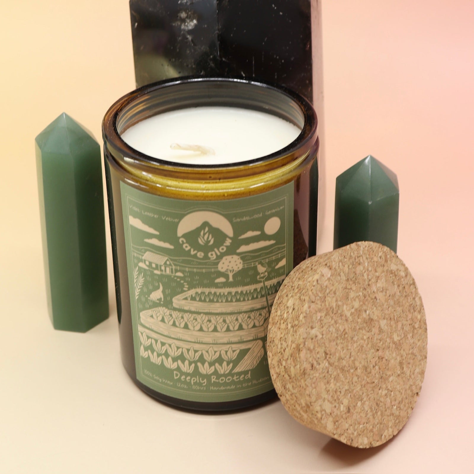 The Deeply Rooted 12oz Soy Candle in front of a large crystal tower surrounded by two smaller crystal towers all on a pink and yellow background.