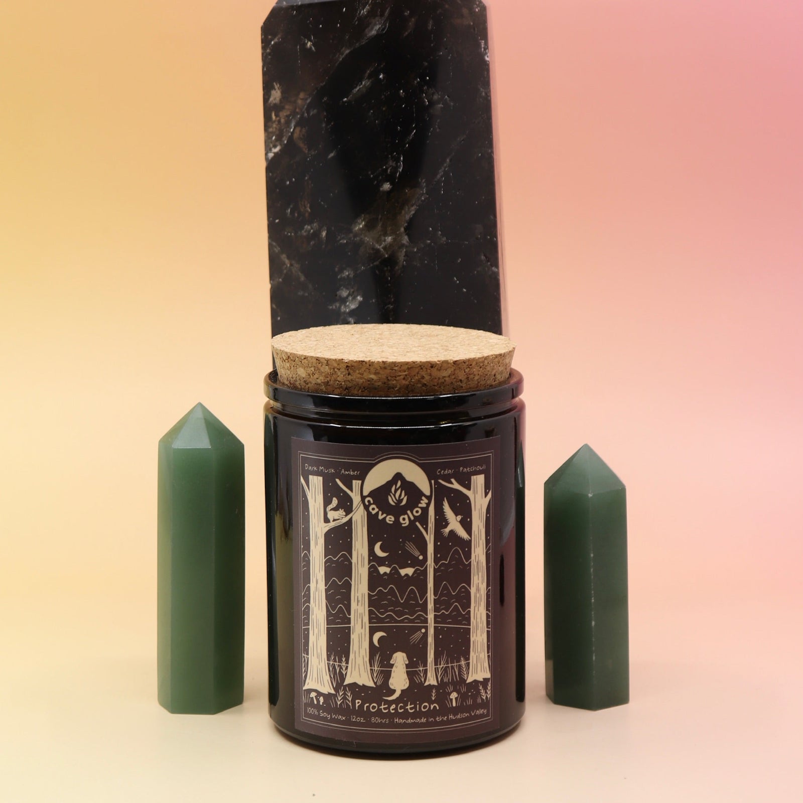 The Protection 12oz Soy Candle in front of a large crystal tower surrounded by two smaller crystals all on a pink and yellow background.