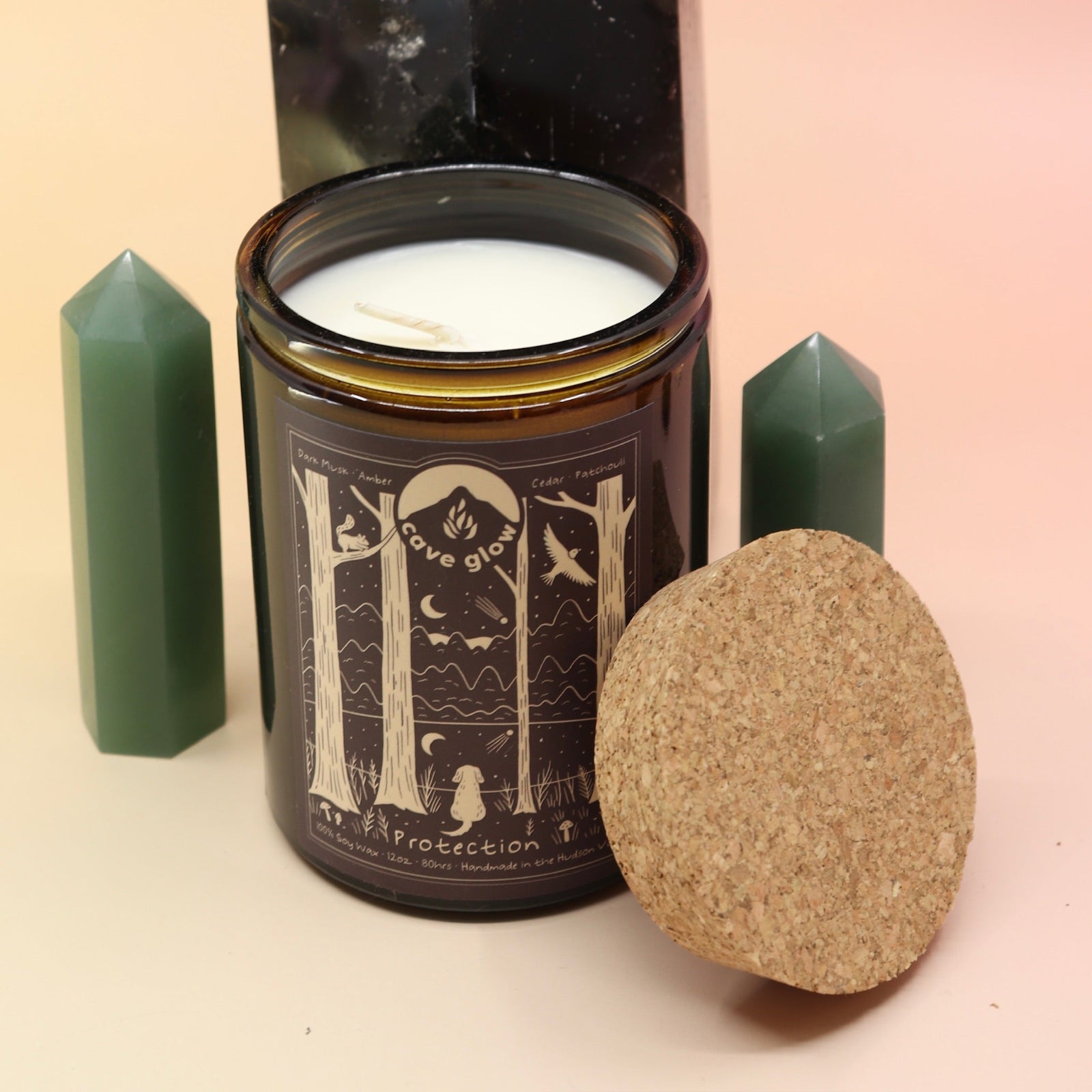 The Protection 12oz Soy Candle in front of a large crystal tower surrounded by two smaller crystals all on a pink and yellow background.