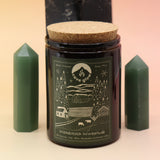 The Woodstock Winterlude 12oz Soy Candle in front of a large crystal tower and two smaller crystal towers on its sides all on a pink and yellow background.