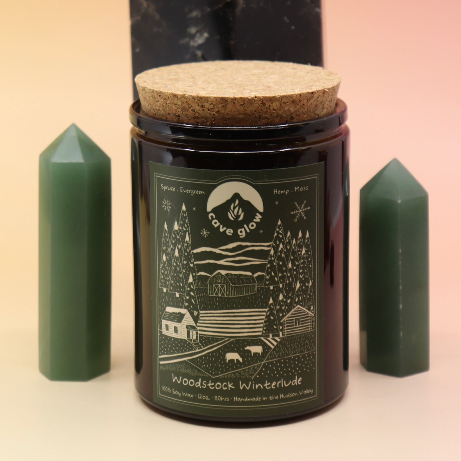 The Woodstock Winterlude 12oz Soy Candle in front of a large crystal tower and two smaller crystal towers on its sides all on a pink and yellow background.