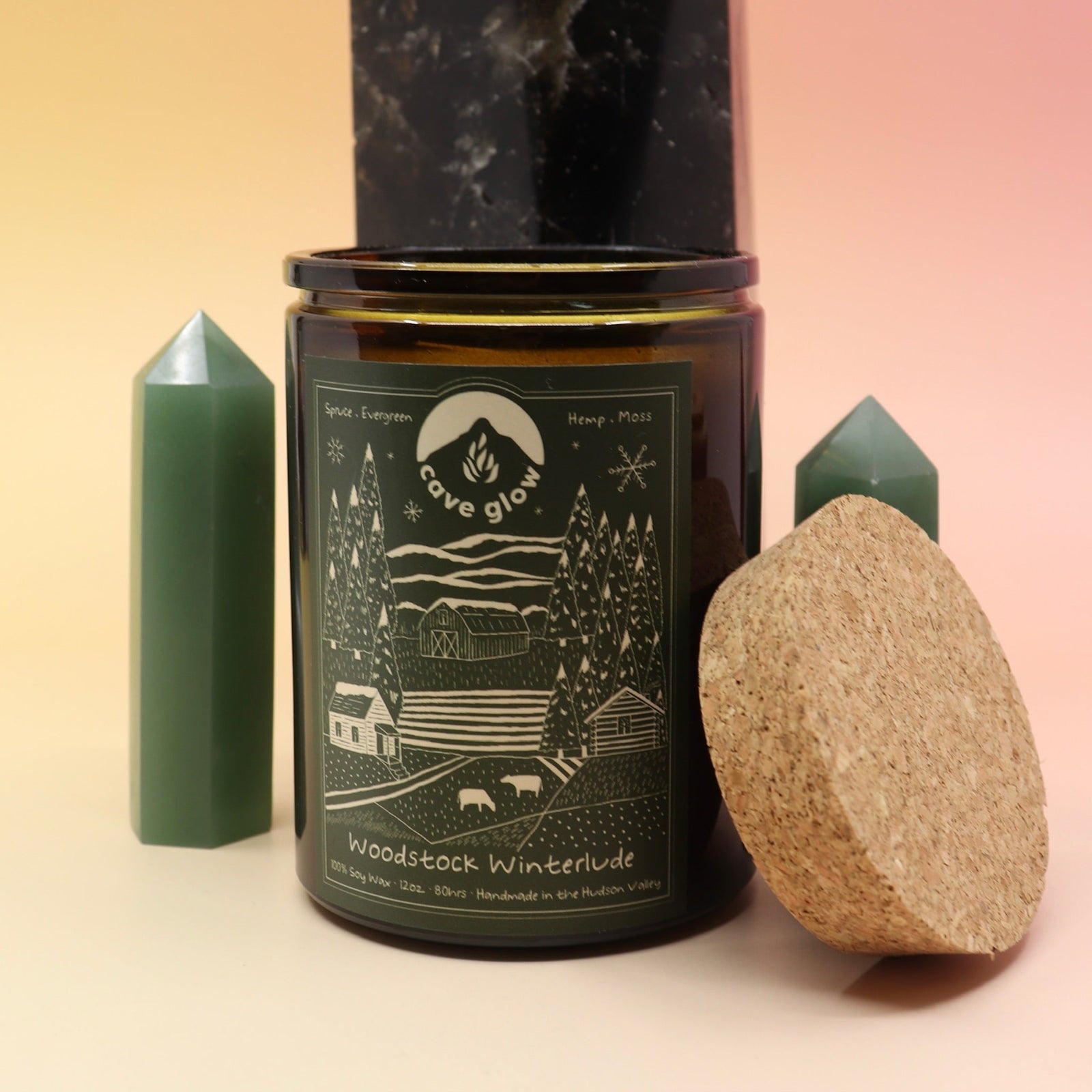 The Woodstock Winterlude 12oz Soy Candle in front of a large crystal tower and two smaller crystal towers on its sides all on a pink and yellow background.