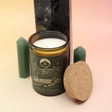 The Woodstock Winterlude 12oz Soy Candle in front of a large crystal tower and two smaller crystal towers on its sides all on a pink and yellow background.