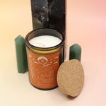 The Poet's Ledge 12oz Soy Candle in front of a large crystal tower and surrounded by two smaller crystal towers all on a pink and yellow background.
