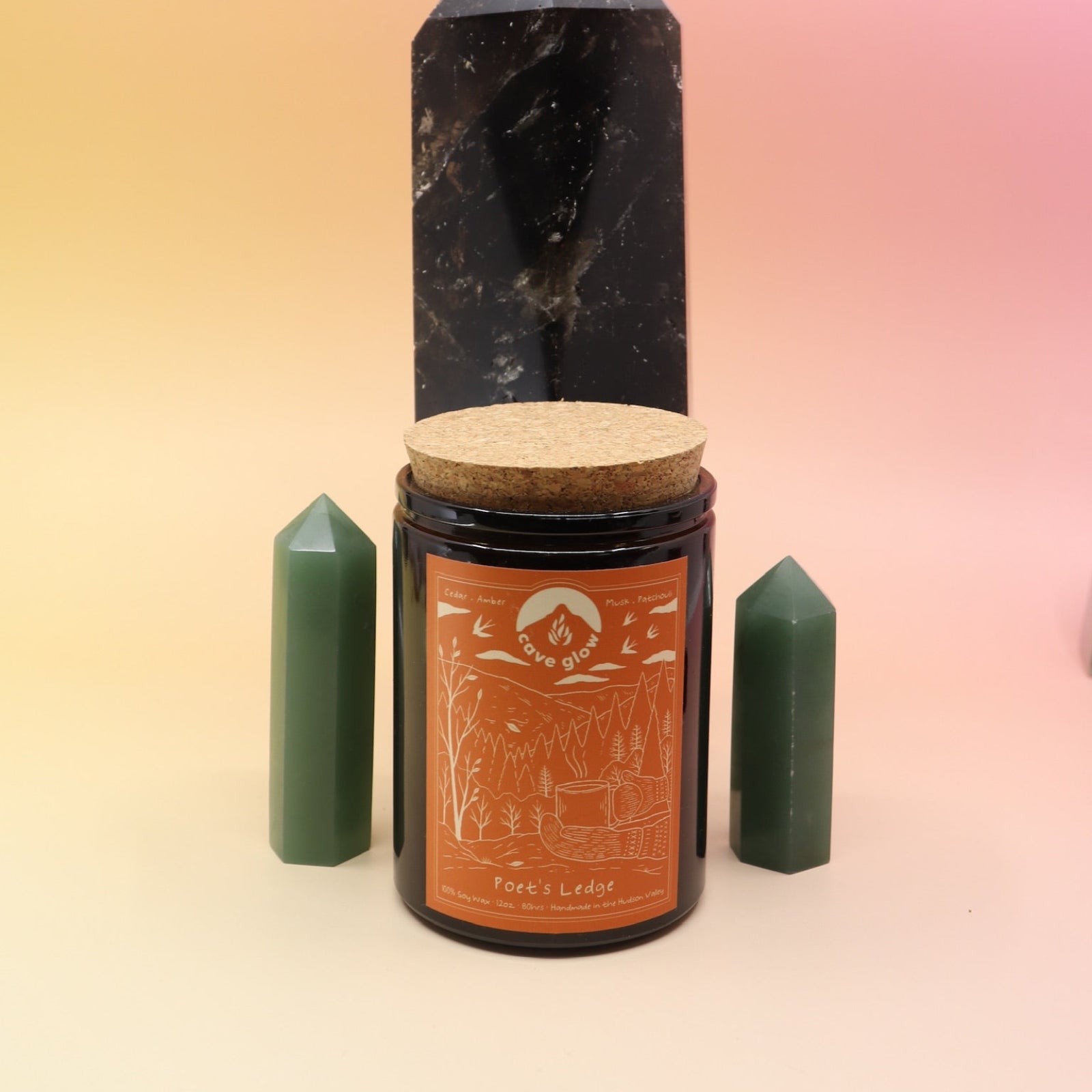 The Poet's Ledge 12oz Soy Candle in front of a large crystal tower and surrounded by two smaller crystal towers all on a pink and yellow background.