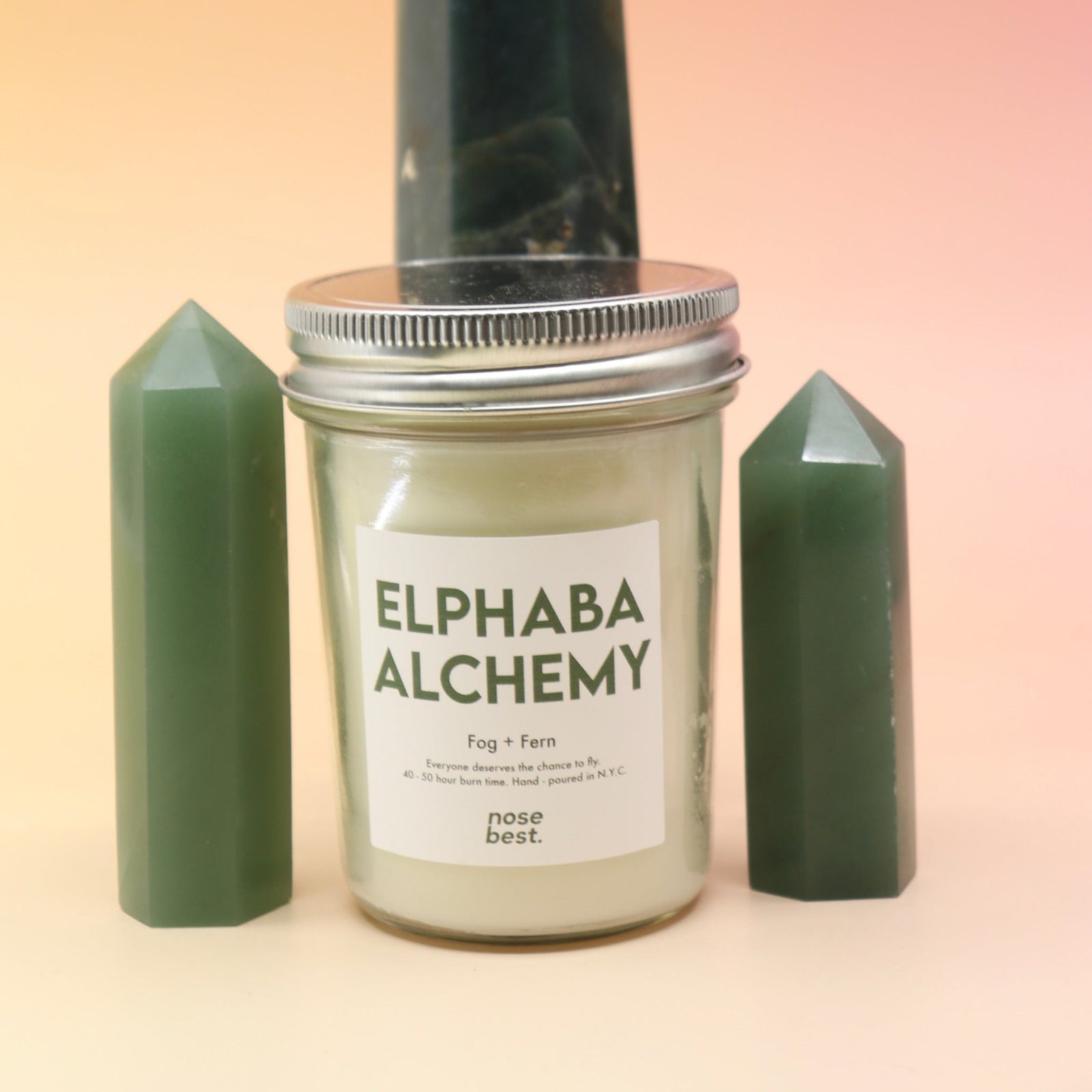 The Fog & Fern Soy Candle surrounded by green crystal tower all on a pink and yellow background.