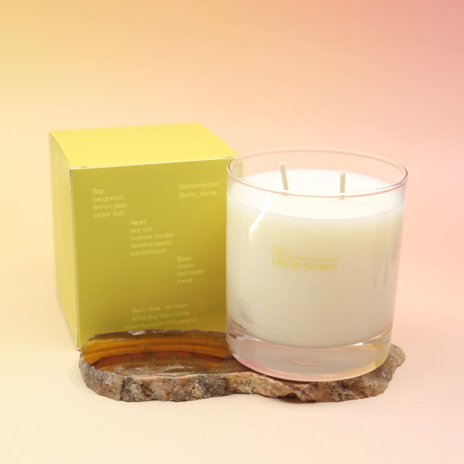 The Hinoki Sesame Candle on an agate slice next to its box all on a pink and yellow background.